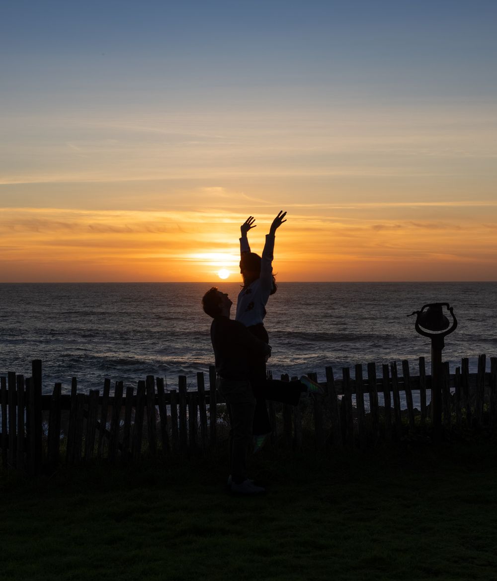 Silhouettes of people and objects against a dramatic sunset over the ocean with a wooden fence in the foreground.