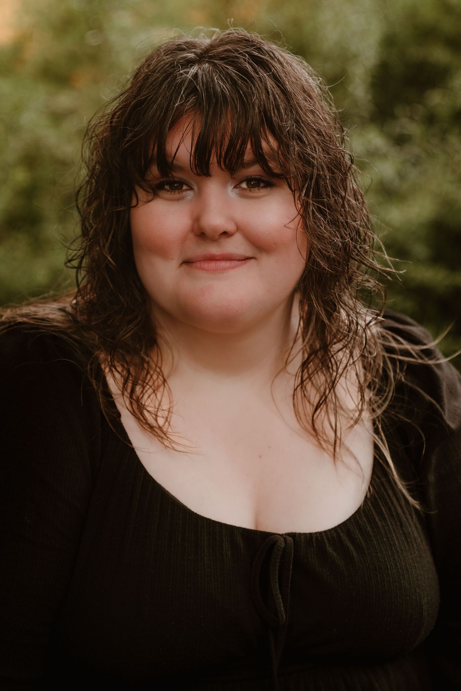Portrait of young woman with dark curly bangs wearing black top, posing outdoors with green foliage background.