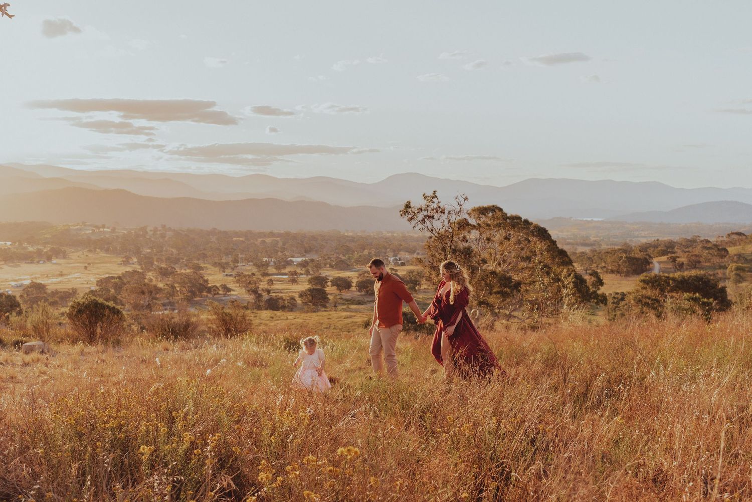 Three people walk through a golden grassland during sunset with mountains visible in the distance.