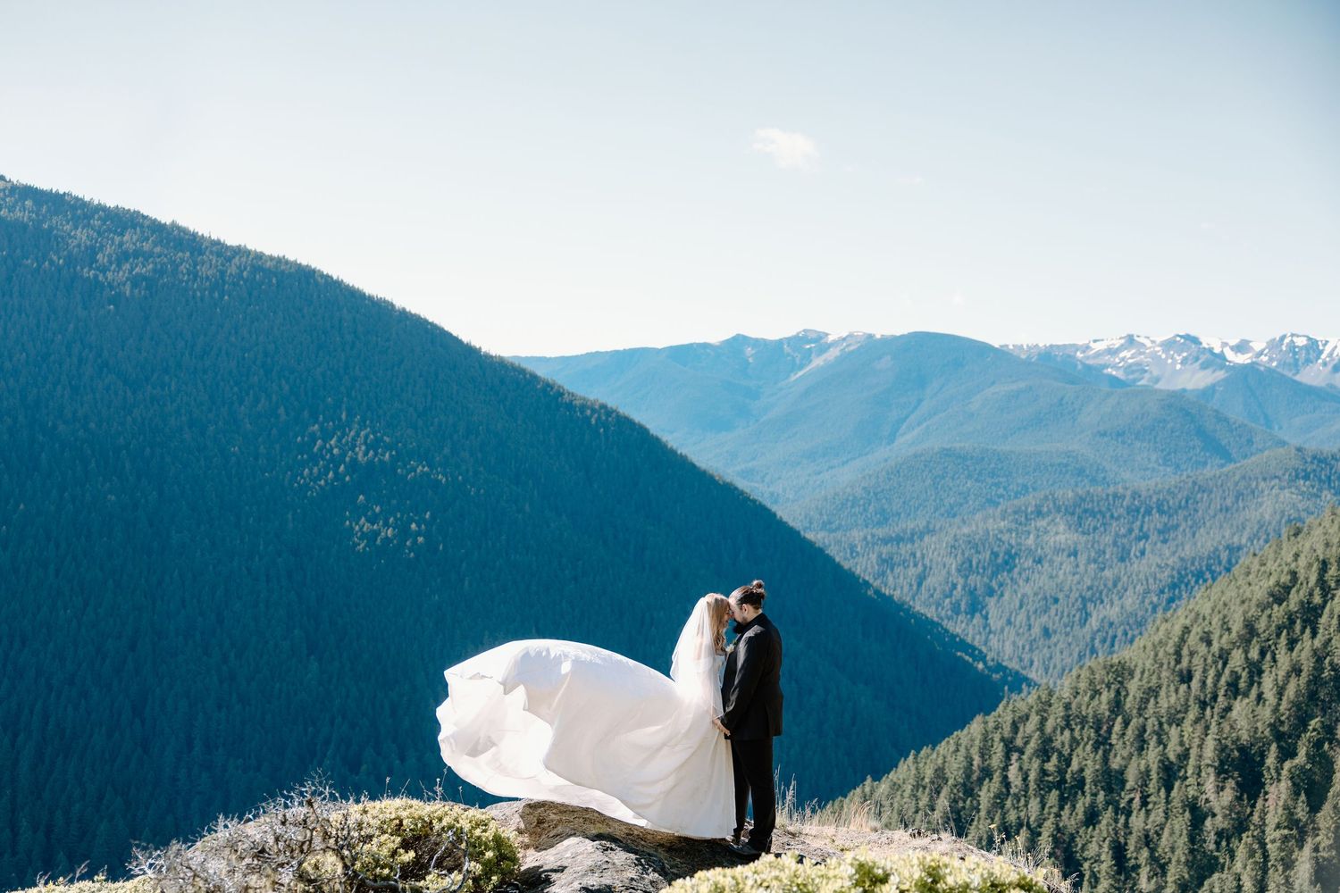 Bride and groom embrace on mountain summit with sweeping forest valley views on sunny wedding day.