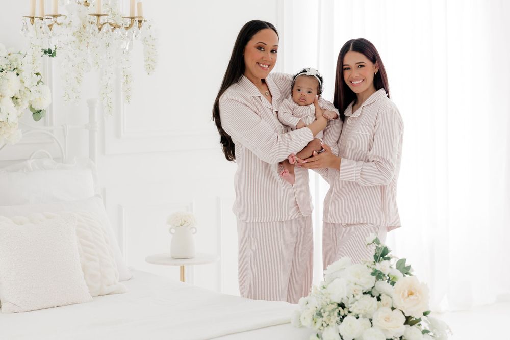 Two people in beige outfits holding a baby in front of an elegant white backdrop with floral decorations.