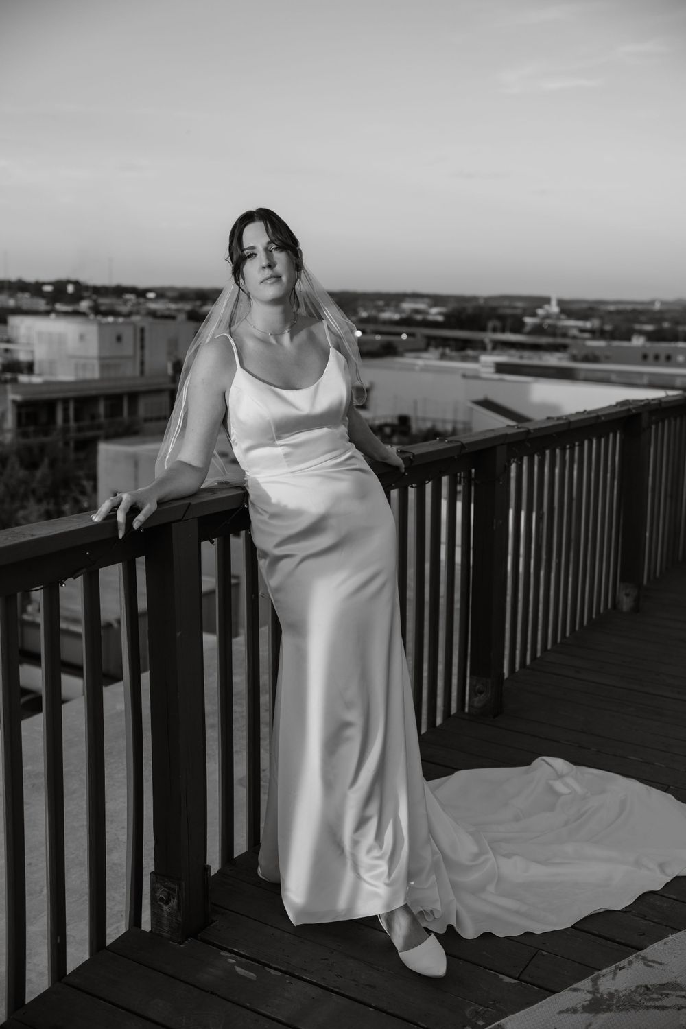 Bride in elegant silk wedding dress poses on balcony overlooking city skyline.