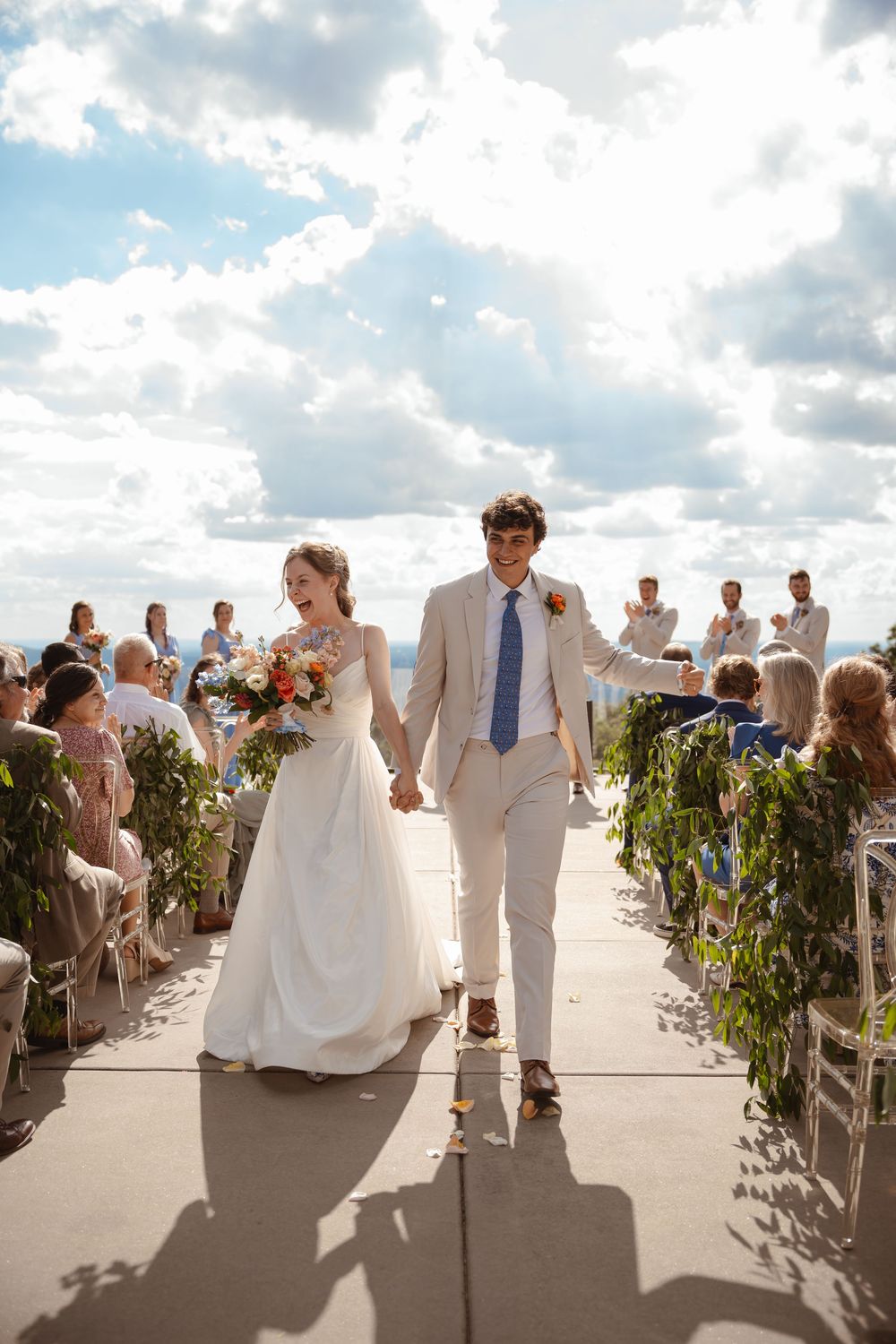 Wedding ceremony processional on a sunny terrace with greenery-lined aisle and white attire.