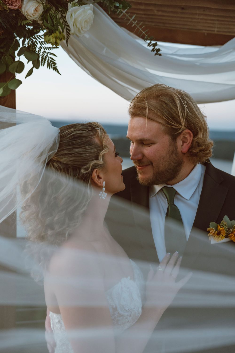 A romantic wedding moment captured through flowing white fabric on an outdoor terrace with floral decorations.
