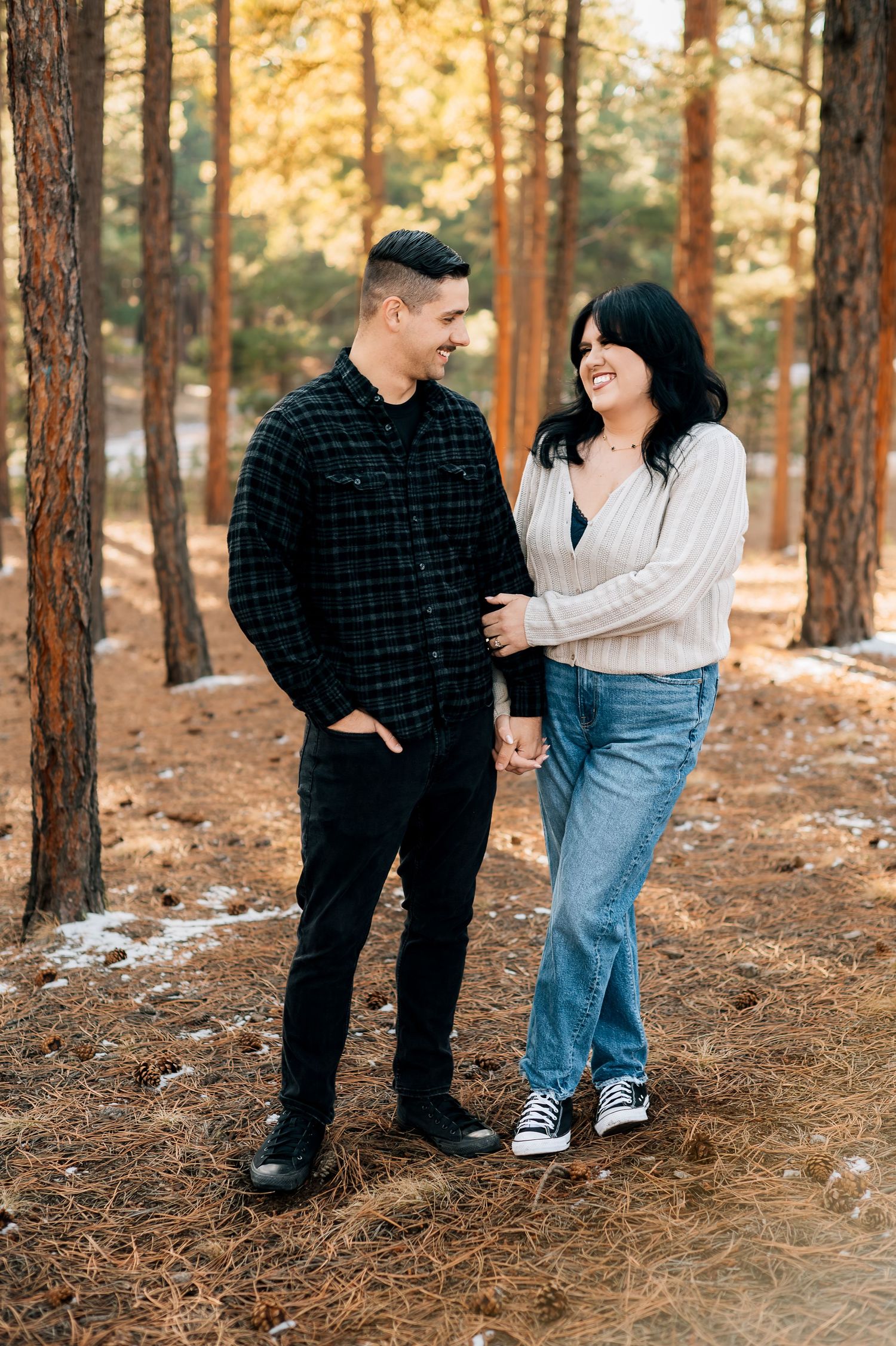 A couple laughs together while standing among tall pine trees in autumn lighting.