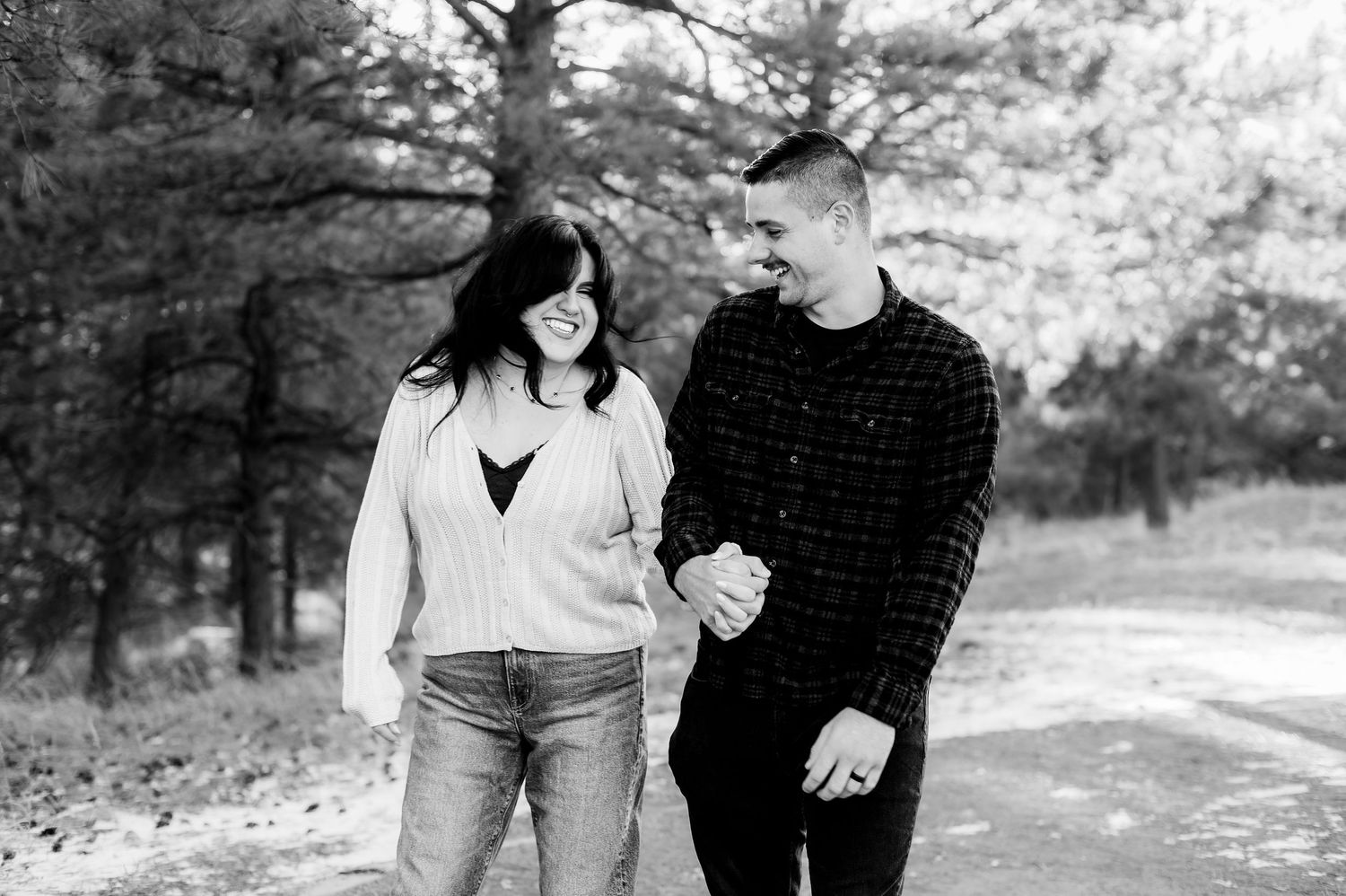 A couple shares a casual moment together in a black and white photo among pine trees.