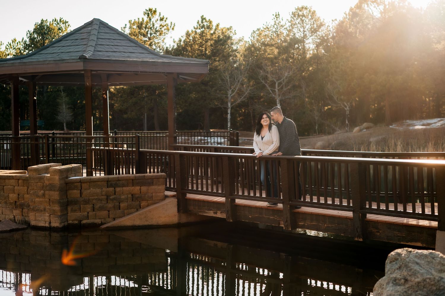 A couple stands on a wooden bridge overlooking water during golden hour.