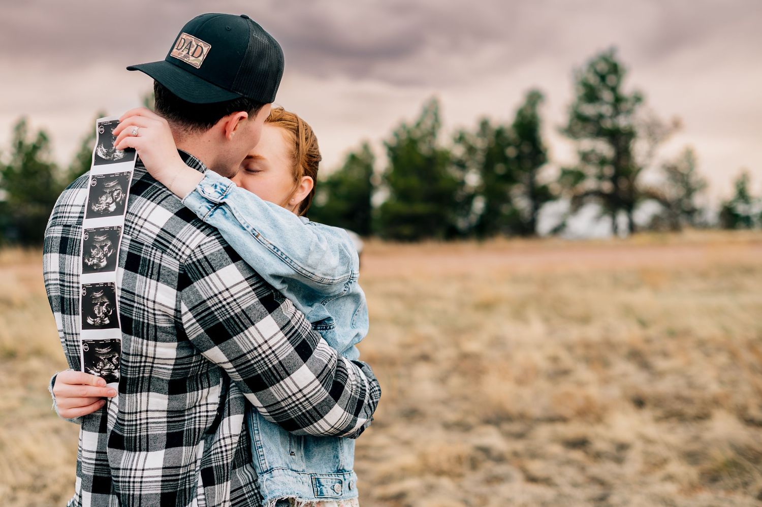 A couple embraces in a plaid shirt and denim against a cloudy rural backdrop.