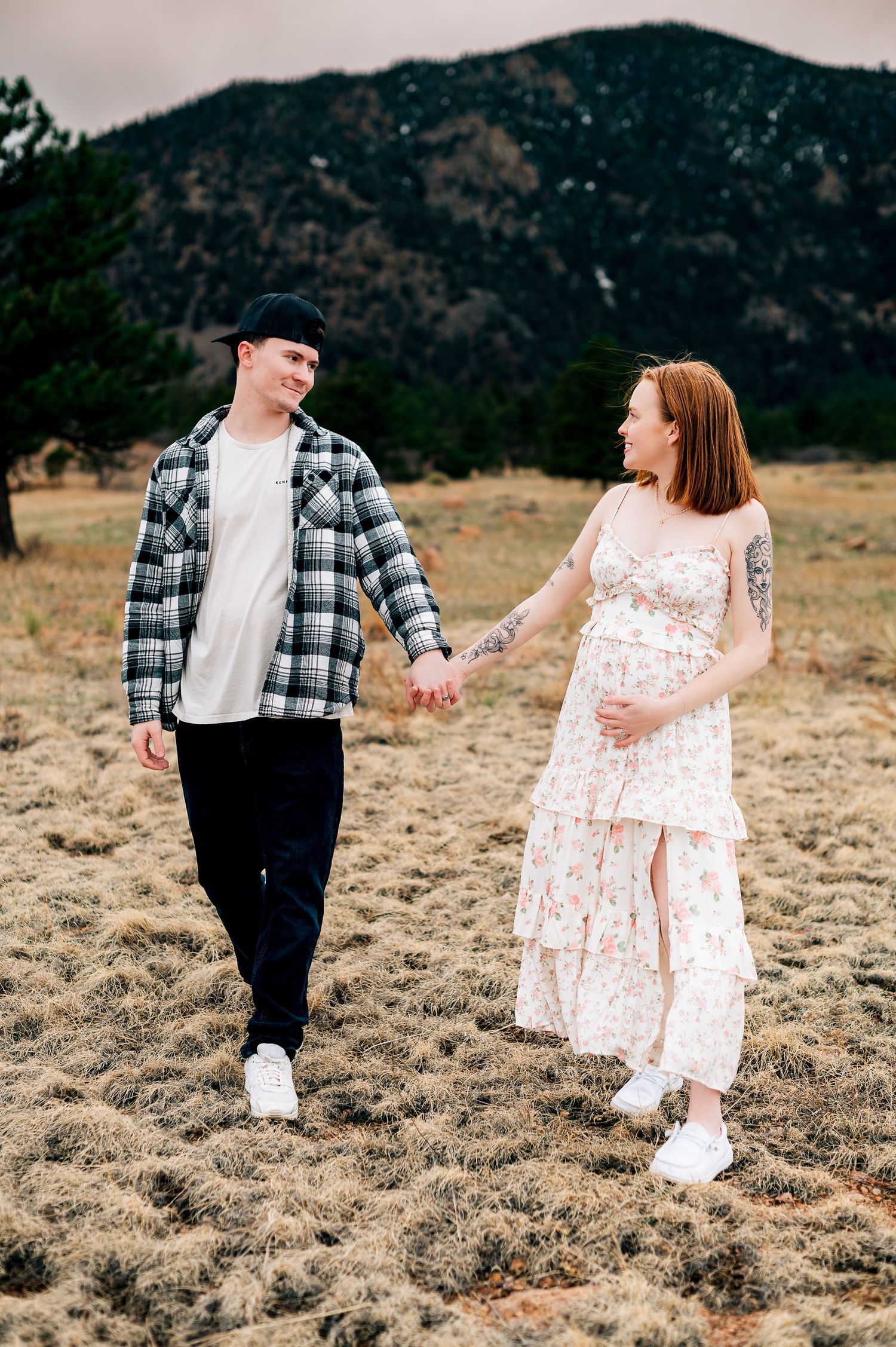 A couple walks hand in hand in a field with mountain views, wearing casual attire.