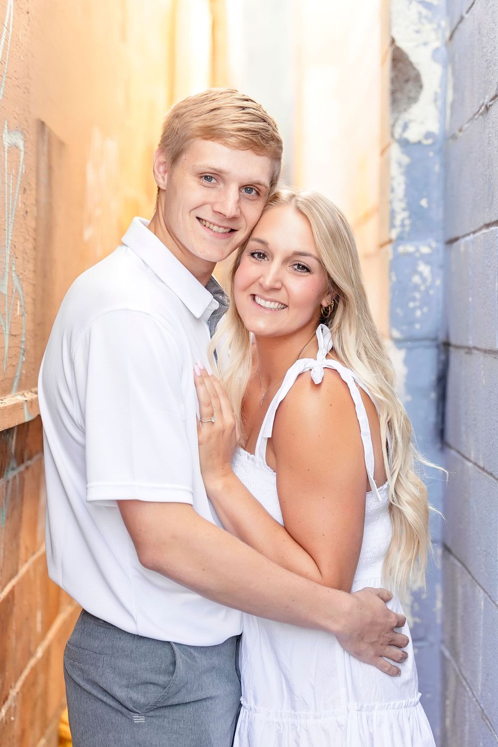 A romantic couple poses against a wall in coordinated white and gray outfits for engagement photos.