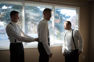 Grooms suspenders are adjusted by his best man, they are backlit by a large window. His second groomsman stands by the window.