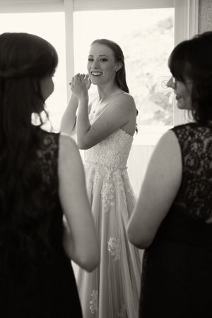 Frames by the window, bride adjusts her earrings as her bridesmaids react to her dress.