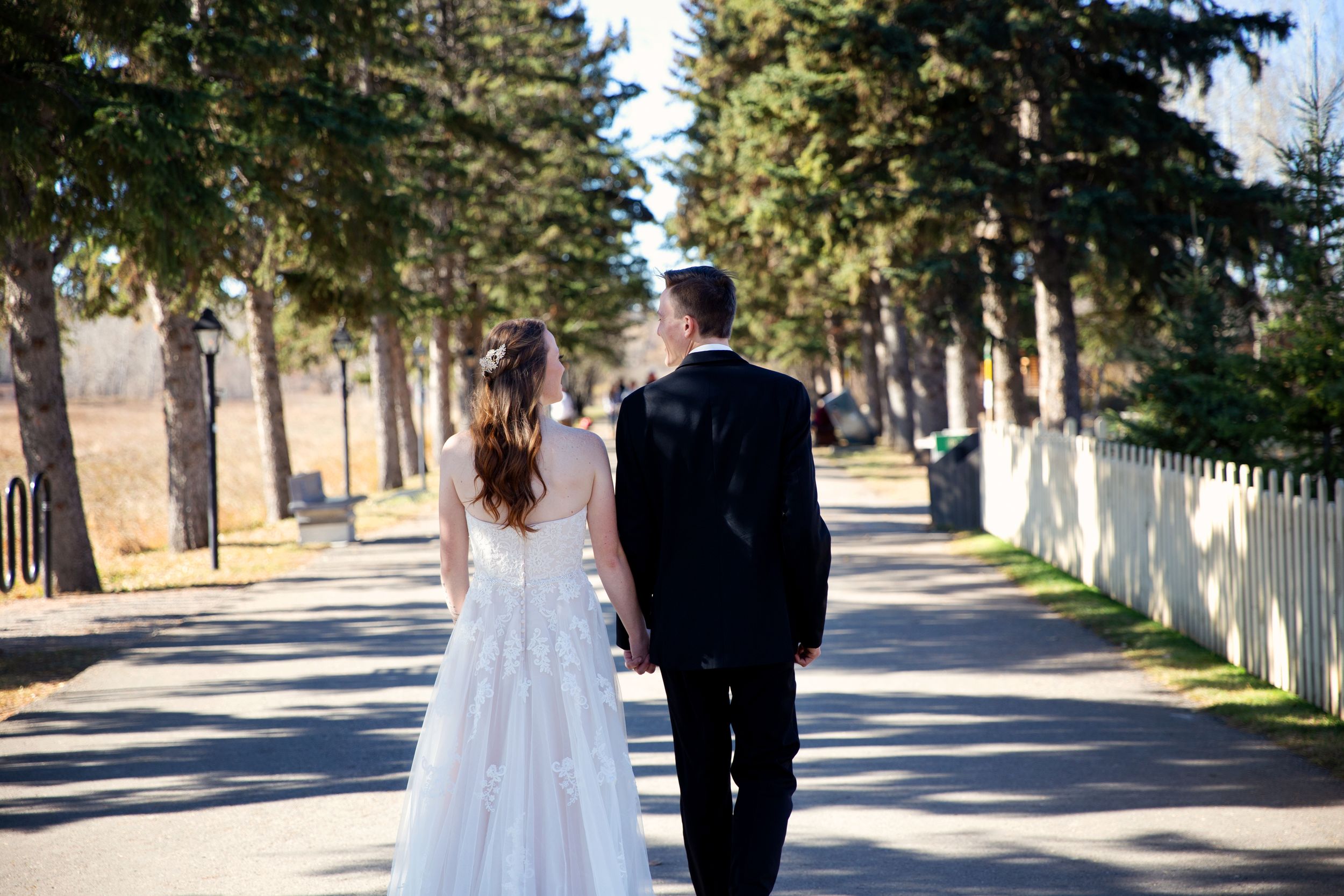 Bride and groom look at each other smiling while walking away from camera down a pathway lined with trees.