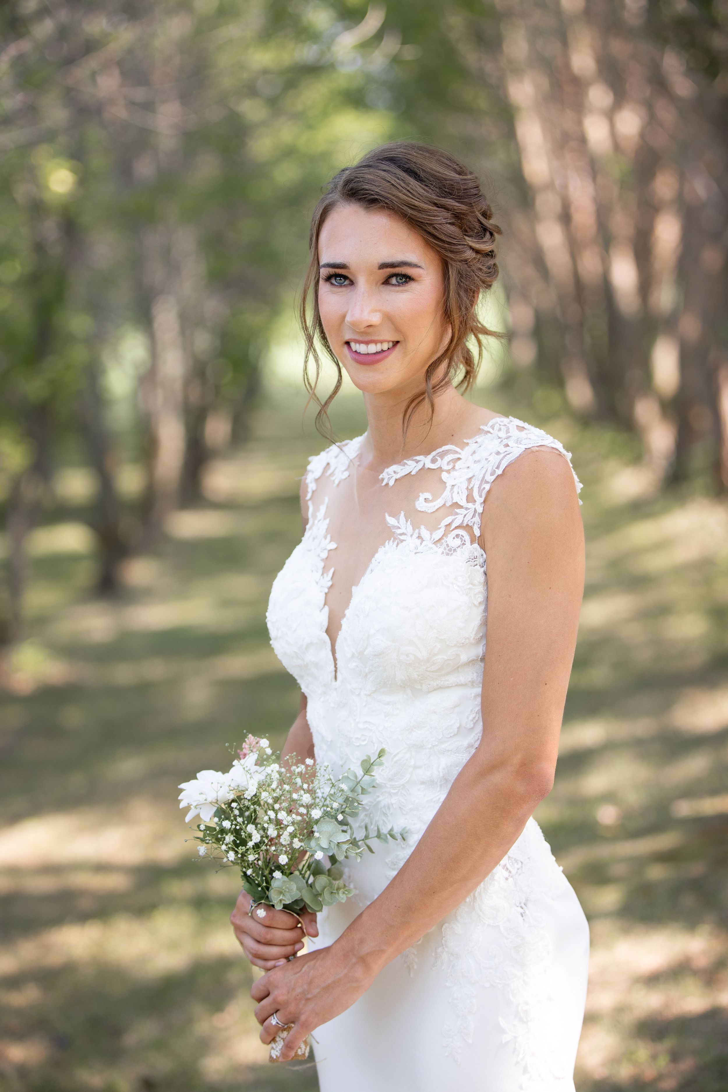 Bride in a white dress stands in a forest of trees holding a small bouquet of white flowers.