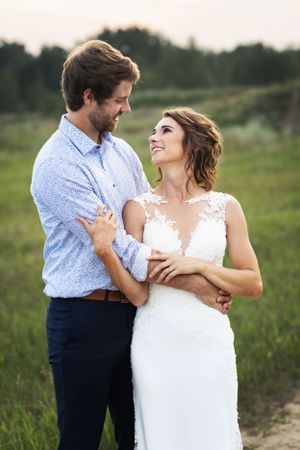 Bride and groom stand together in a green field arms wrapped around each other.