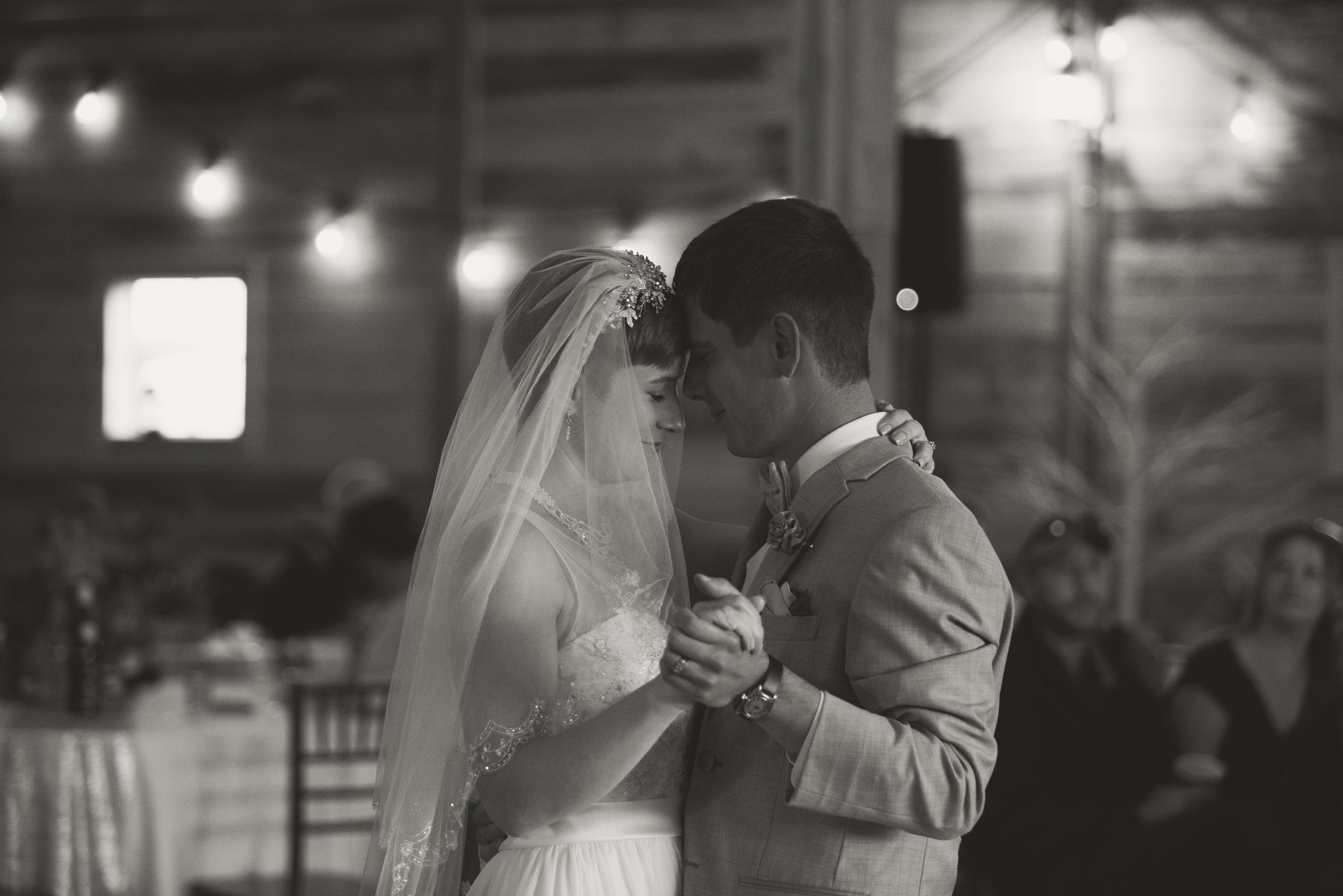 Black and white photo of bride and groom sharing their first dance. Twinkling lights scattered in the background.