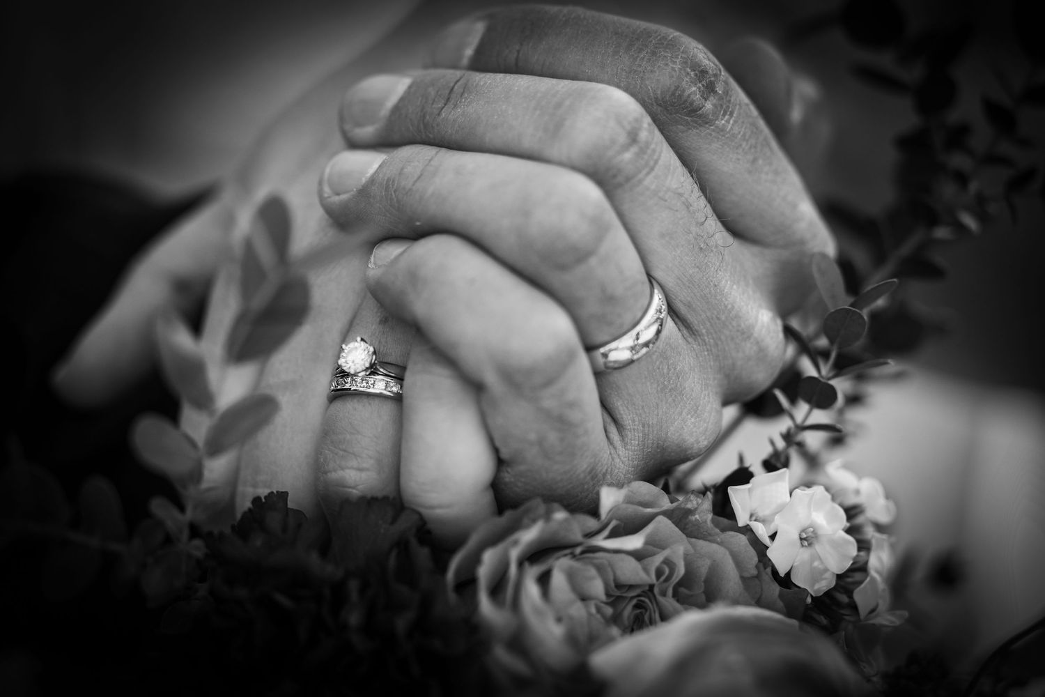 Black and white close-up of clasped hands with wedding rings among flowers.