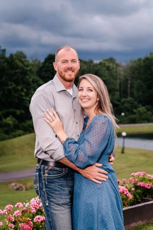 Couple posing together in casual attire near a pond with stormy clouds overhead.