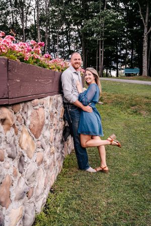 Couple embracing by stone wall with pink flowers in casual summer outfits.
