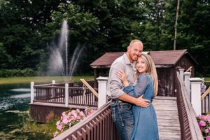 A couple embraces on a wooden deck overlooking a pond with a fountain and pink flowers.
