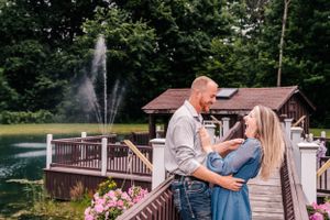 A couple embraces on a wooden walkway with a scenic pond and fountain in the background.