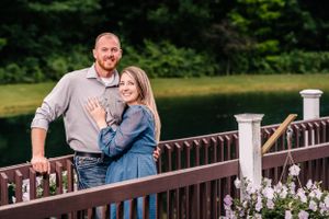 A happy couple poses on a wooden bridge with flowering plants and railing.