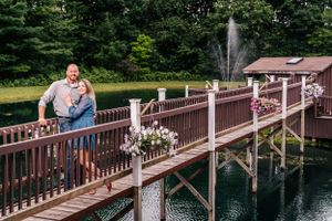 A couple stands on a wooden bridge over a pond with a fountain in the distance.
