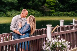 A romantic couple shares an intimate moment on a wooden bridge with white railings and flowers.