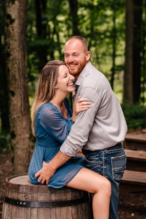A couple shares a joyful embrace while sitting on a rustic wooden barrel in a forest setting.