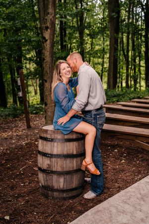 A couple poses together on a wooden barrel in a wooded outdoor setting.