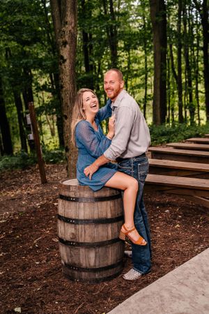 A couple laughing together while sitting on a rustic barrel in a forest setting.