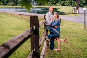 A couple poses playfully against a wooden fence with a lake in the background.