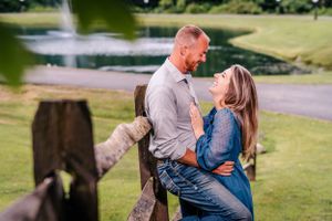 A couple shares a romantic moment by a rustic wooden fence with a fountain in the background.