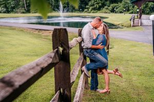 A couple embraces by a wooden fence near a pond with a water fountain.