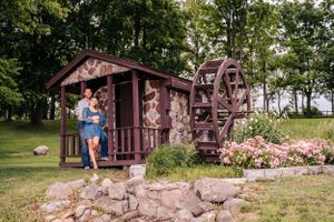 A rustic wooden cabin with a waterwheel surrounded by pink flowers and greenery.