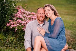 An engaged couple poses in front of pink flowering bushes during a summer photo session.