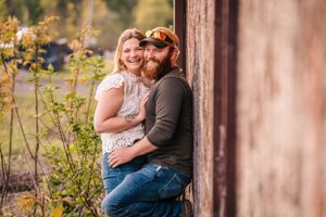 A couple embraces while leaning against a rustic wall surrounded by autumn foliage.