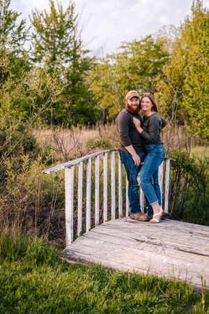 A couple poses together on a white wooden footbridge in a natural setting.