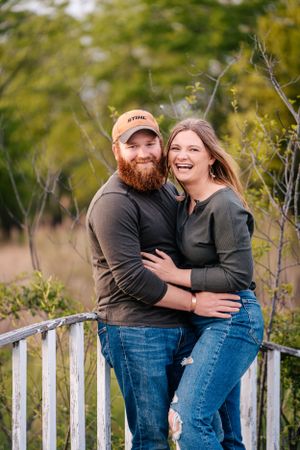 A happy couple embraces while standing on a white wooden bridge surrounded by greenery.
