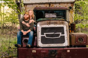 A couple poses on an old rustic truck in a wooded setting.
