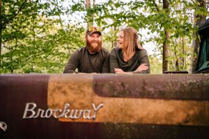 A relaxed couple poses on an antique Brockway truck in a forest setting.