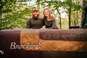A casual portrait of a couple on a rustic Brockway truck surrounded by trees.
