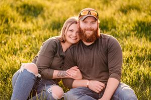 A couple sits together in a golden field at sunset wearing casual brown clothing.