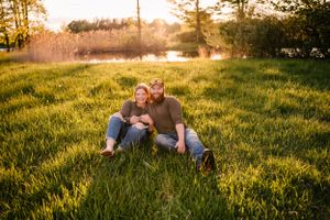 A couple relaxes on lush grass during golden hour with sunlit trees in the background.