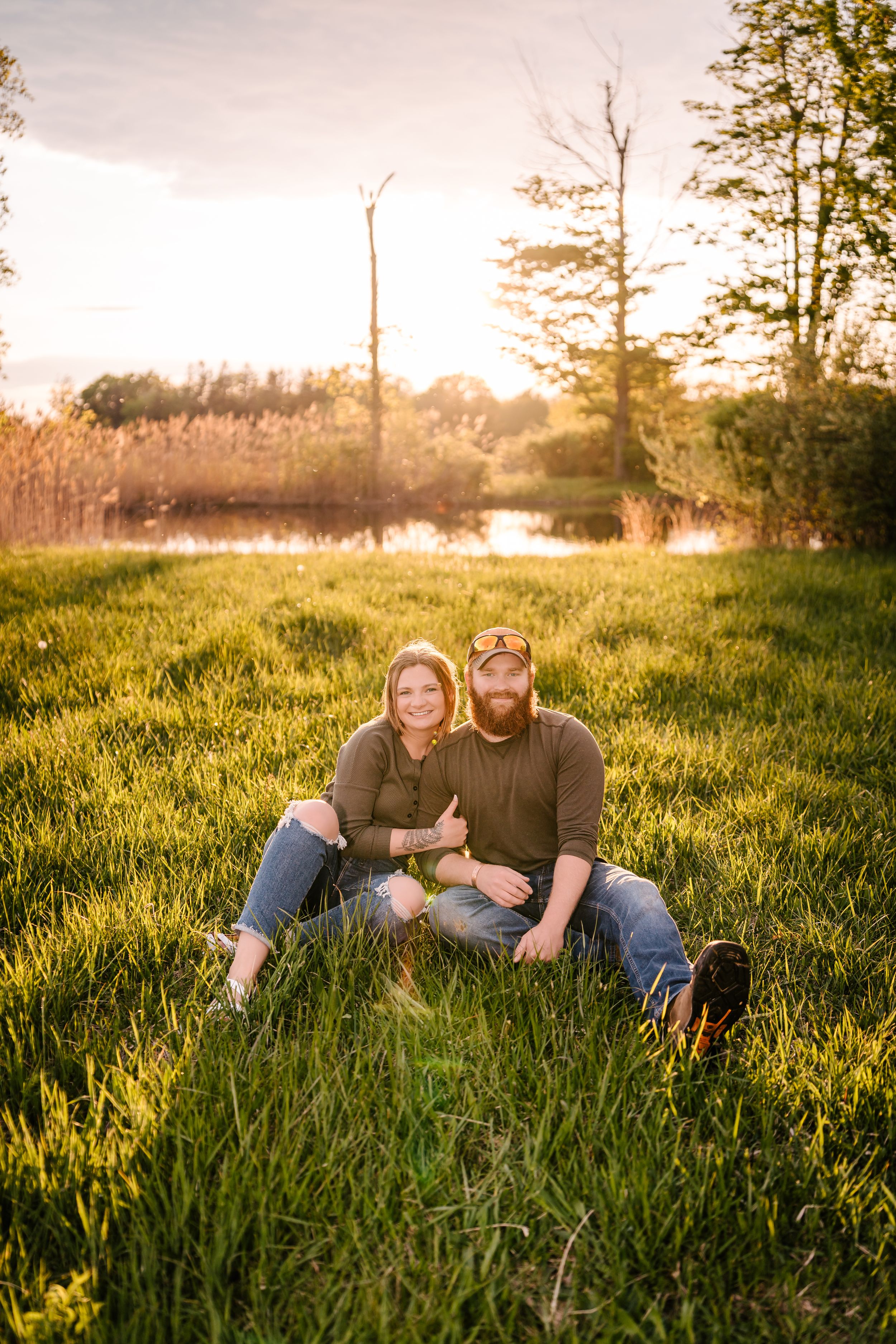 A couple poses together on grass near a pond during sunset with trees in the background.