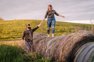 Two people balance and play on large round hay bales in a rural field at sunset.