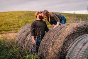 Two people playfully interact on large round hay bales in a grassy field.