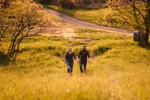 Two people walk together on a winding dirt path through golden grass at sunset.