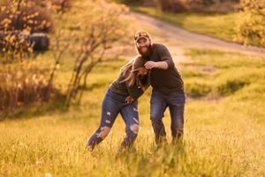 Two people playfully wrestle in tall golden grass during sunset.