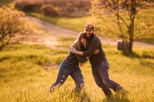 Two people embrace while playing in a sunlit field with a dirt path nearby.